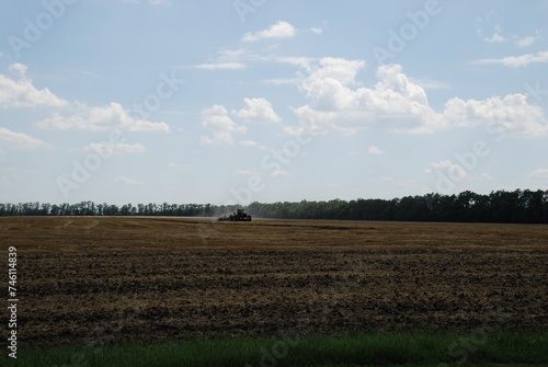 A mown field under sky. Summer sunny day in the village. A wide field in which yellow, withered, mown stems of plants and black soil remained. A tractor drives across the field and digs up the soil.