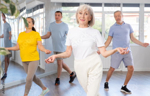 Fototapeta Naklejka Na Ścianę i Meble -  Positive elderly woman practicing Tai Chi with group of aged people, promoting health and wellness in bright training room