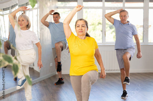 Positive elderly woman practicing Tai Chi with group of aged people, promoting health and wellness in bright training room