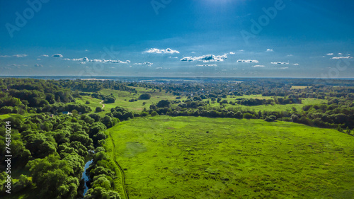 Vast, flat terrain covered with green arable fields and meadows. You can see a dirt road, clumps of trees, and a coniferous forest on the outskirts. View from height, photo taken with a drone.