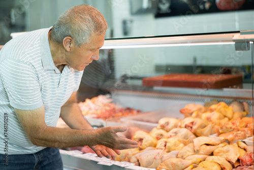 Interested elderly man looking at large assortment of fresh raw meat products in glass refrigerated display case in butchery shop