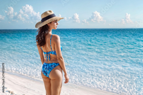 Back view of young woman in swimsuit and straw hat on tropical beach. A woman in a bikini stands on a beach.