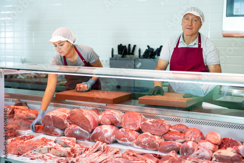 In organic natural meat store, friendly farmer male seller stands near counter refrigerator display case. In butcher shop, elderly male vendor in uniform apron, gloves, cap holds cutting board