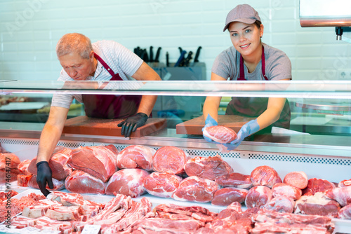 Young female seller offers shows piece of veal meat fillet for cooking family barbecue dinner. Mature male sales assistant packs order in background