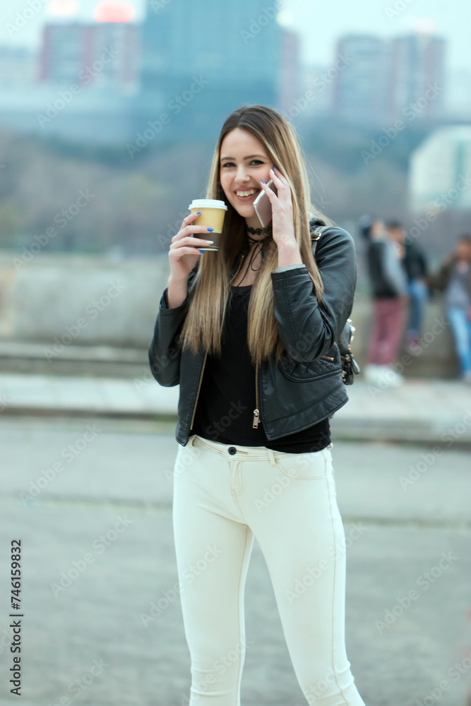 Young woman on the phone with a coffee. Dreamy hipster girl enjoying leisure time while walking at spring urban setting with gadget