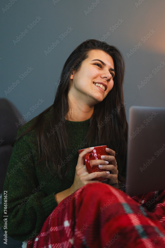 A young girl or woman getting up from bed drinking coffee or tea in blanket cozy room while using laptop for work and talk with friends or family on video call	