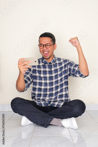 Adult Asian man sitting crossed leg showing excited when looking to cellphone photo