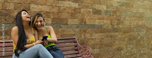 Two multiracial girls laughing watching media content on line in their smart phones sitting on a bench background bricks yellow brown friendships concept
