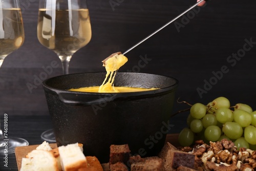 Dipping piece of bread into fondue pot with melted cheese on table  closeup