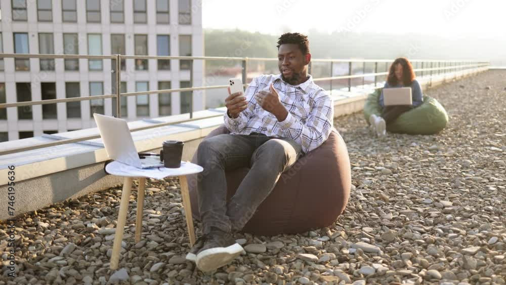 Joyful African man in casual checkered shirt using phone while recording video for her vlog on panoramic terrace. Smiling blogger demonstrating city view for subscribers on sunny day outdoors. Joyful African man in casual checkered shirt using phone while recording video for her vlog on panoramic terrace. Smiling blogger demonstrating city view for subscribers on sunny day outdoors.