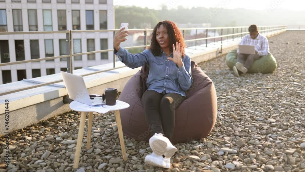 Joyful African woman in casual denim shirt using phone while recording video for her vlog on panoramic terrace, while male colleague working on blurred background. Joyful African woman in casual denim shirt using phone while recording video for her vlog on panoramic terrace, while male colleague working on blurred background.