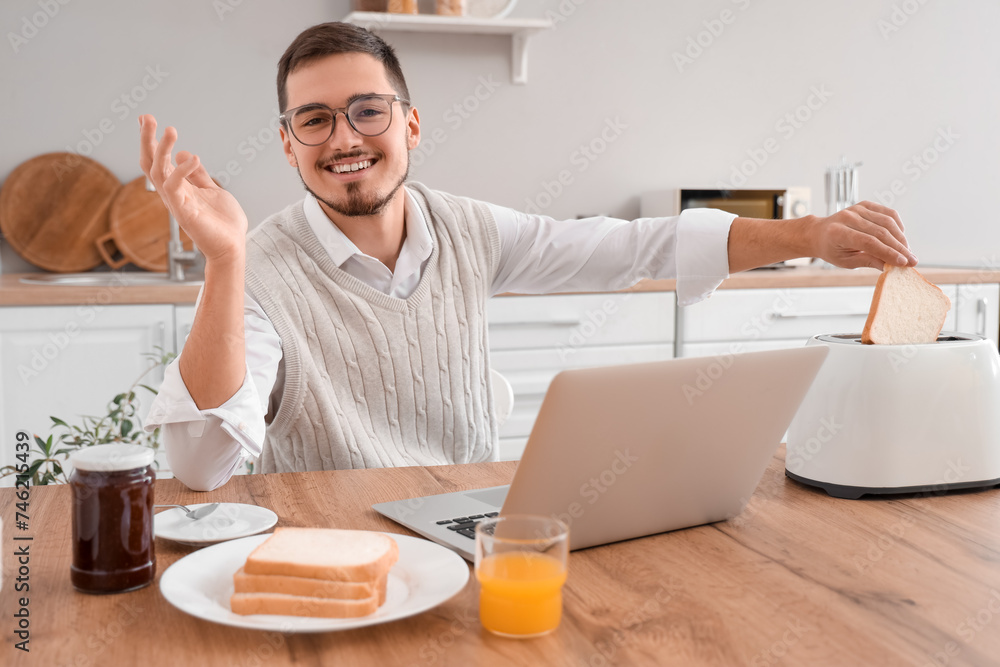 Young man with laptop having tasty toasts for breakfast in kitchen Young man with laptop having tasty toasts for breakfast in kitchen