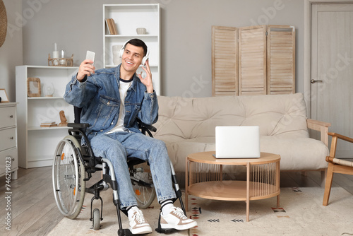 Happy young man in wheelchair with headphones using mobile phone at home
