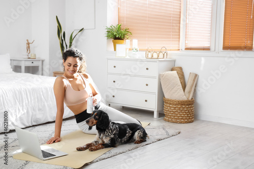 Sporty young African-American woman with cocker spaniel and laptop in bedroom