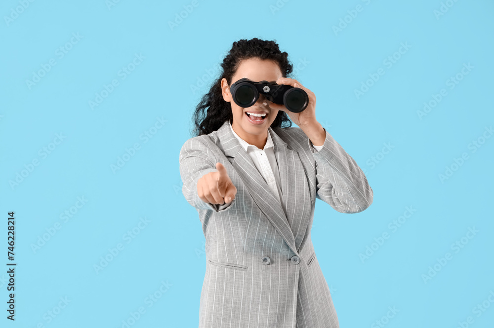 Young African-American businesswoman with binoculars pointing at viewer on blue background Young African-American businesswoman with binoculars pointing at viewer on blue background