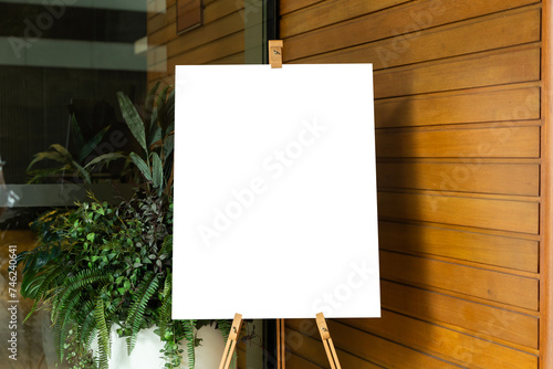 A blank white canvas sits on an artist's easel in a studio. A potted plant stands next to the easel. The canvas is pristine and white.