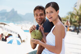 Couple, coconut and drink at beach with smile for vacation with milk, cocktail and juice in portrait. People, man and woman with sunshine, review and fruit for water, happy and detox with nutrition