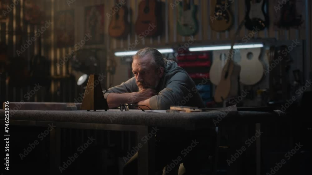 Tired artisan, craftsman looks at metronome sitting in stylish workshop for making musical instruments. Mature carpenter rests after long workday. Wooden guitars hang on the wall. Handmade concept. Tired artisan, craftsman looks at metronome sitting in stylish workshop for making musical instruments. Mature carpenter rests after long workday. Wooden guitars hang on the wall. Handmade concept.