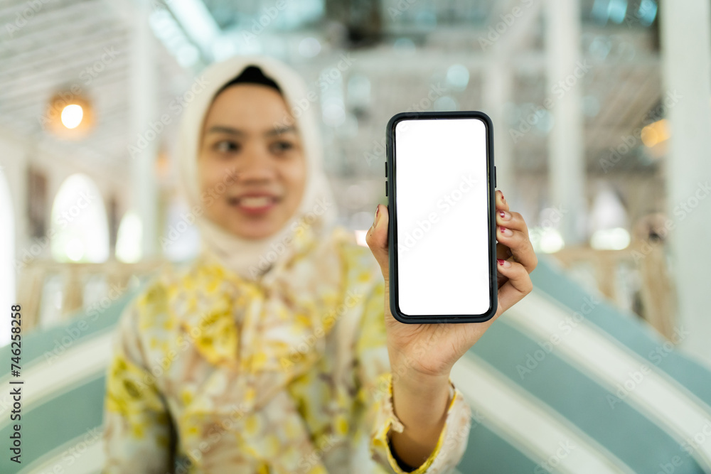 Beautiful Asian woman, wearing a casual hijab pointing to the smartphone screen. Sit back and relax in a cafe or restaurant.
