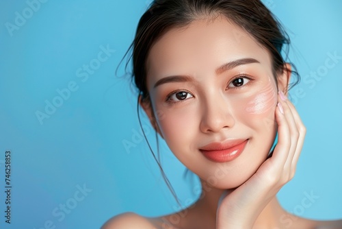 A beautiful smiling Asian model girl with natural makeup touches radiant moisturized skin on a blue background in close-up. The concept of beauty treatments and skin care.
