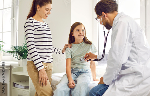 Mother and child come together to their family doctor. Pediatrician or family practitioner puts stethoscope to chest of happy little girl and listens to her lungs or heartbeat. Medical exam concept  photo