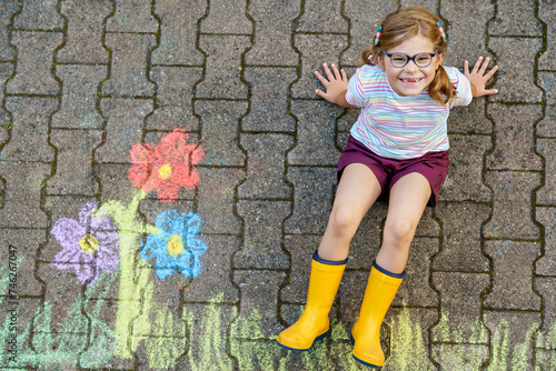 Cute little girl and flowers painted with colorful chalks on asphalt. Happy preschool child having fun with painting chalk picture. Creative leisure for children, drawing and painting.