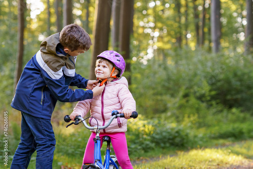 School kid boy, brother put on little preschool sister girl bike helmet on head. Brother teaching happy child cycling and having fun with learning bike. Active siblings family outdoors. Kids activity