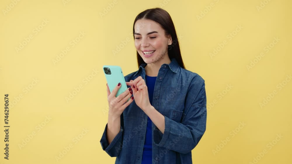 Dark haired woman with happy smile on face standing over yellow background and tapping on screen of modern smartphone. Caucasian female in casual clothes chatting online during pastime in studio.