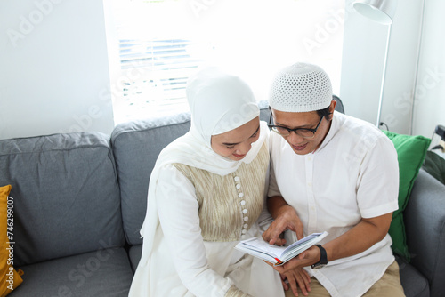 Asian Muslim man teaching woman reading Koran or Quran in living room. Muslim couple praying on the couch at home.