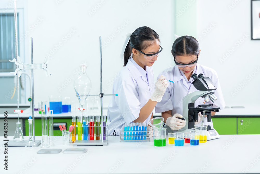 Two female Asian students are scientists at the medical faculty. Drop the test solution into the microscope. Produce and research drugs to treat viral diseases There are beakers in the biology lab. Two female Asian students are scientists at the medical faculty. Drop the test solution into the microscope. Produce and research drugs to treat viral diseases There are beakers in the biology lab.
