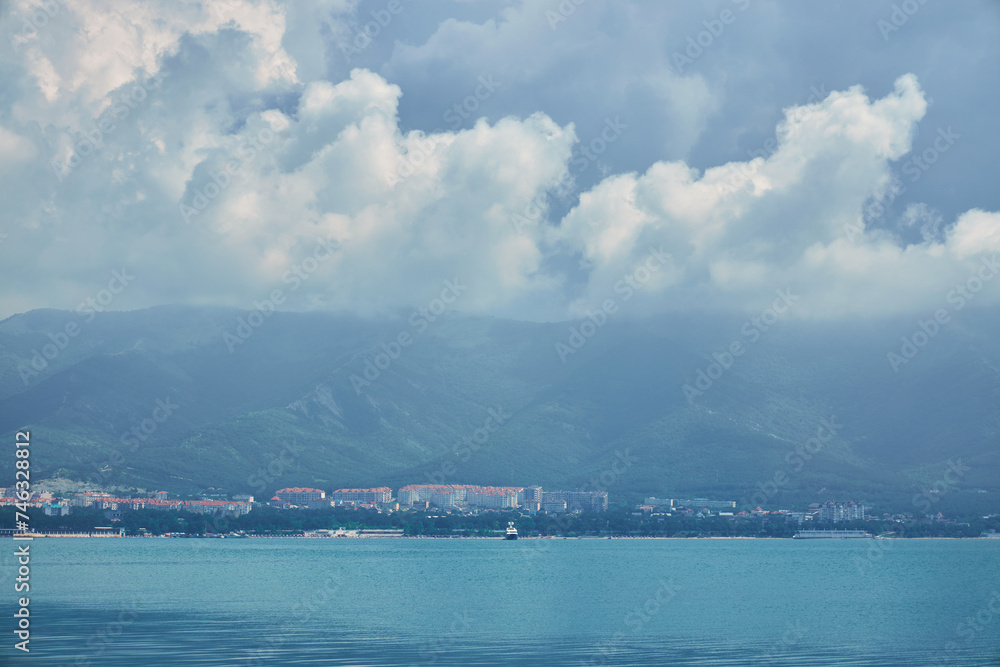 Coastal city nestles at the foot of misty mountains, with the calm sea in the foreground and a sky filled with clouds