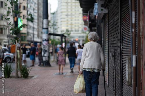 A grandmother returning home after shopping, carrying a bag and a cane.