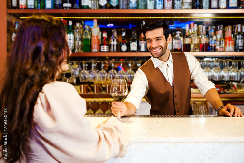 Caucasian professional bartender or mixologist making a cocktail for women at a bar. An attractive barman served a glass of wine with a smile to a woman. Bartender service at the night club restaurant