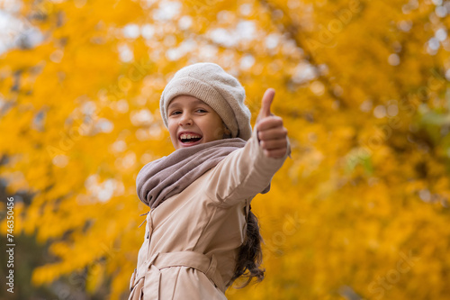 Happy caucasian girl in a beige coat and beret walks in the park in autumn. Schoolgirl showing thumbs up. 