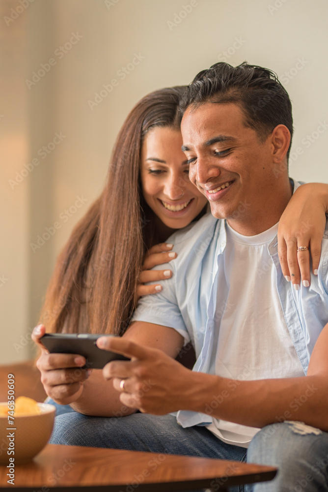 Modern young couple at home sitting on the sofa playing with mobile phone games app together having fun and enjoying indoor leisure activity together. Couple man woman young in apartment lifestyle Modern young couple at home sitting on the sofa playing with mobile phone games app together having fun and enjoying indoor leisure activity together. Couple man woman young in apartment lifestyle