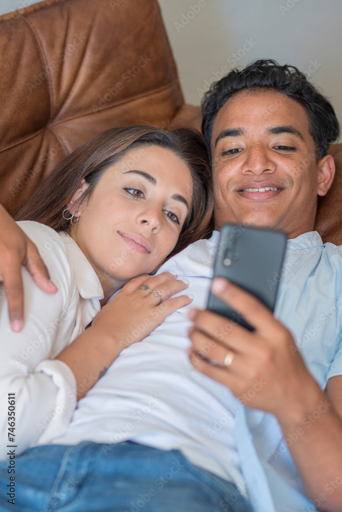Young couple having relax together using mobile phone laying on the sofa in indoor relaxation leisure activity together. Black boy and caucasian girl in love and relationship, Intimate people enjoying Young couple having relax together using mobile phone laying on the sofa in indoor relaxation leisure activity together. Black boy and caucasian girl in love and relationship, Intimate people enjoying