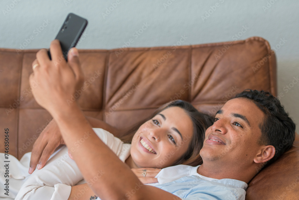 Happy young couple laying on the sofa at home and have fun using mobile phone to take selfie picture for social media pages account. Modern people man and woman living together in relationship love Happy young couple laying on the sofa at home and have fun using mobile phone to take selfie picture for social media pages account. Modern people man and woman living together in relationship love