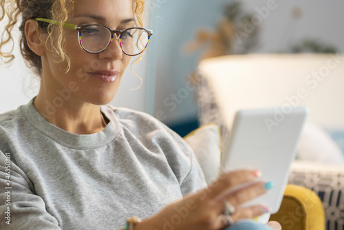 Attractive middle age woman portrait at home reading book on ereader modern device witting comfy on the yellow sofa. Female people in relax indoor leisure activity alone using technology smart tablet photo