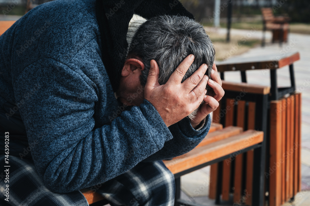 Upset lonely homeless elderly old Caucasian man with depression holds head in hands while sitting on a park bench in autumn Upset lonely homeless elderly old Caucasian man with depression holds head in hands while sitting on a park bench in autumn