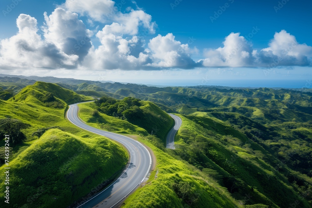 Aerial view of an empty mountain highway weaving through lush green hills on a sunny day, with vibrant blue skies and fluffy white clouds.