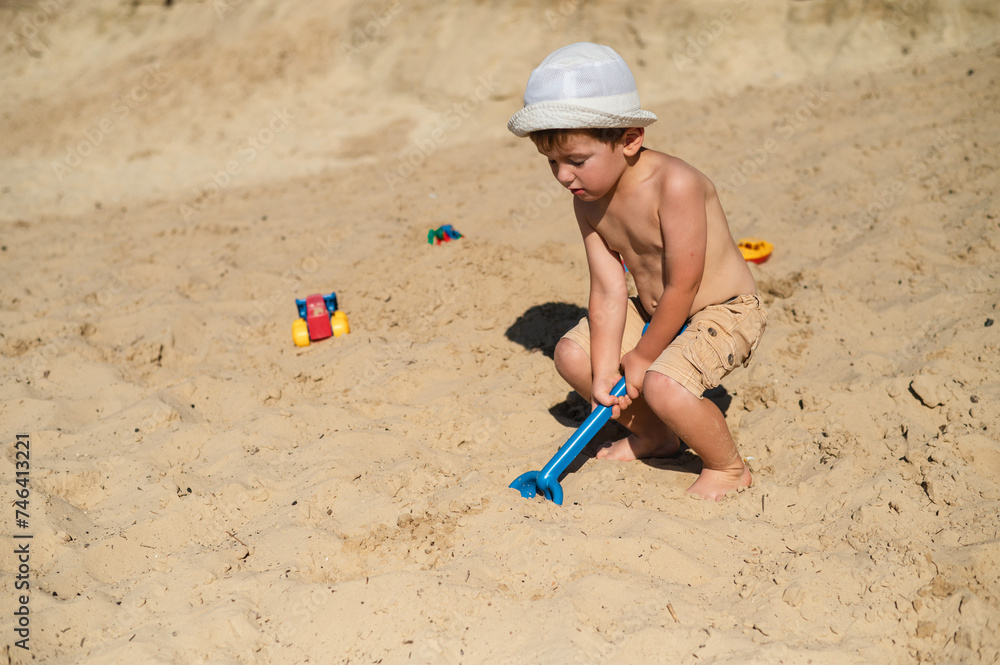 A 3-year-old child builds a sand castle on the beach. Games on the beach on a hot summer day. A boy plays with a stove and makes little beads, pours sand into a bucket with a plastic scoop A 3-year-old child builds a sand castle on the beach. Games on the beach on a hot summer day. A boy plays with a stove and makes little beads, pours sand into a bucket with a plastic scoop