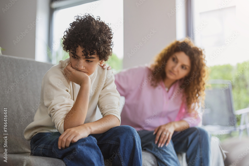 Obraz premium Upset teenage boy sitting on sofa while his mother looking at him