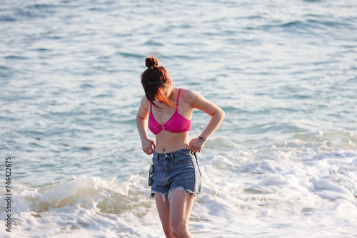 Asian girl in jeans shorts and pink bikini standing on sea beach on surf waves background © Oleg