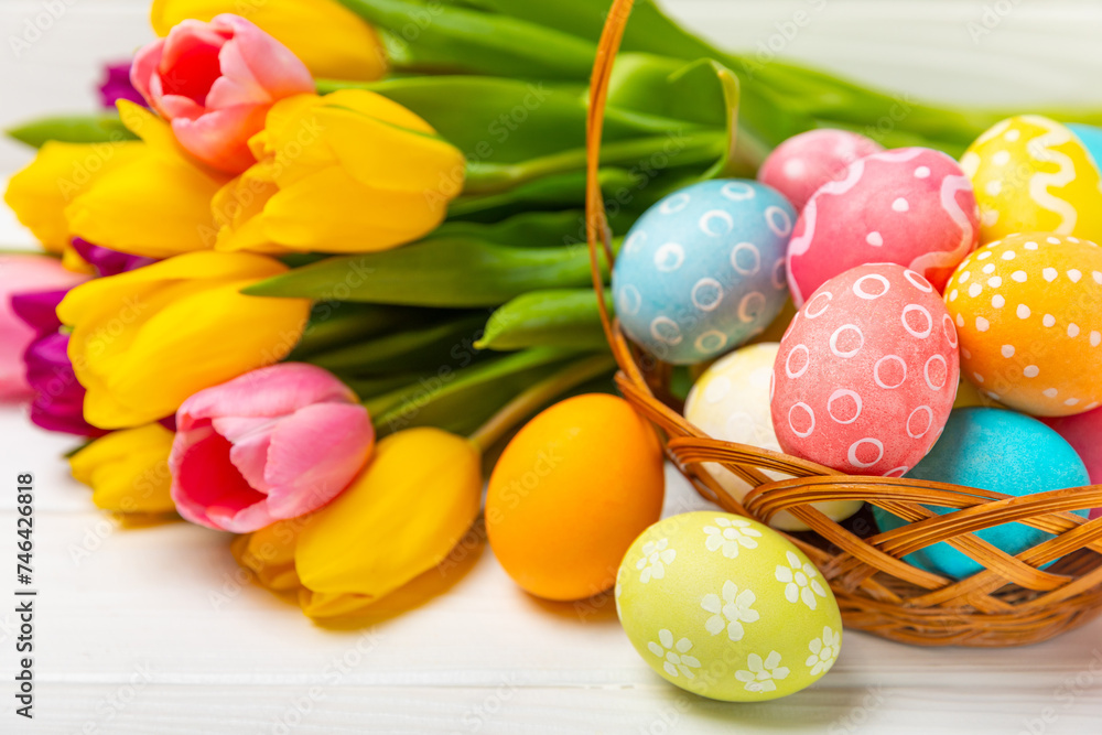 Easter basket filled with colorful eggs and a bouquet of tulips on a textured wooden table. Easter celebration concept. Colorful easter handmade decorated Easter eggs. Place for text. Copy space Easter basket filled with colorful eggs and a bouquet of tulips on a textured wooden table. Easter celebration concept. Colorful easter handmade decorated Easter eggs. Place for text. Copy space
