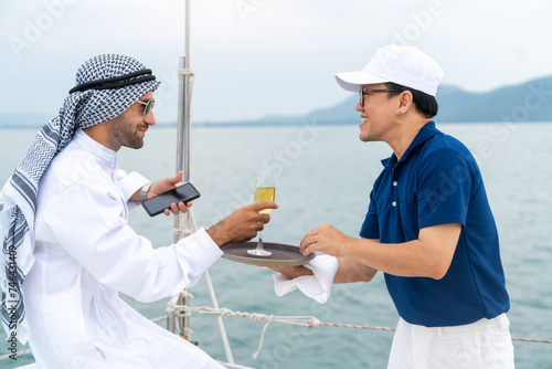 Asian man waiter serving drink to Arab businessman passenger while luxury catamaran boat yacht sailing in the ocean. Arabic man relax and enjoy outdoor lifestyle travel on summer vacation.
