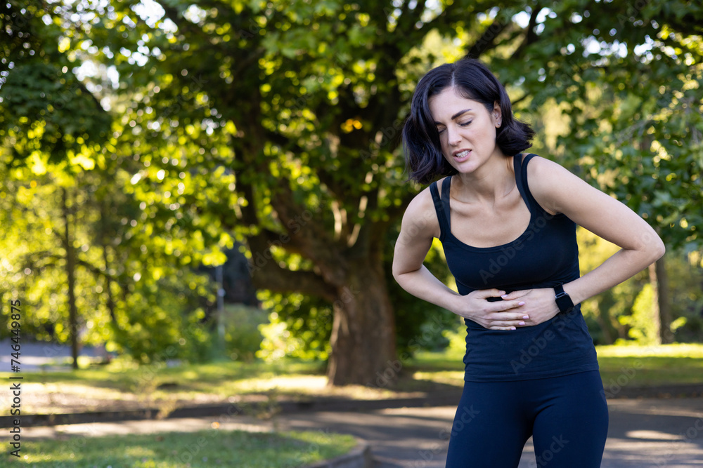 A young woman is standing in the park outdoors, bent over and holding her stomach, feeling severe pain after running and doing sports A young woman is standing in the park outdoors, bent over and holding her stomach, feeling severe pain after running and doing sports