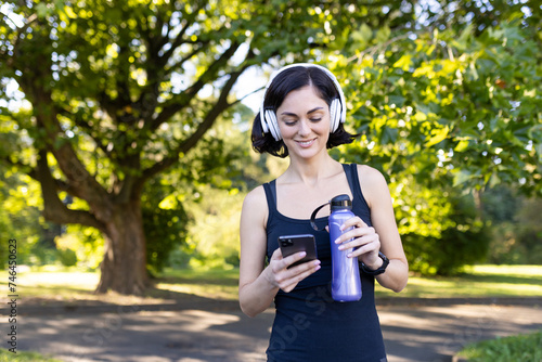 Smiling young woman doing sports and jogging outdoors in park, standing resting in headphones, holding water bottle and looking at phone screen