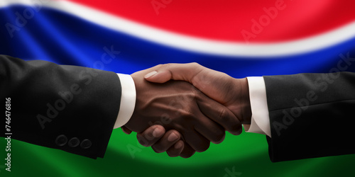 Businessman and diplomat in suits clasp hands for handshake over Gambia flag, agree on united success in trade, diplomacy, cooperation, negotiation, support, teamwork in commerce, gesture of greeting