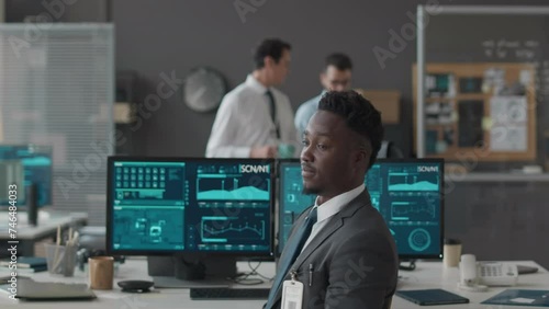 Medium slowmo portrait of young African American male cybersecurity officer turning to camera on office chair and posing for camera, sitting in front of multiple computer displays at office desk photo