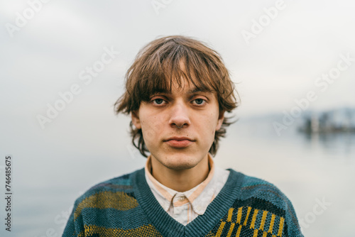 Fototapeta Young male with contemplative gaze, wearing sweater, against blurred lakeside backdrop